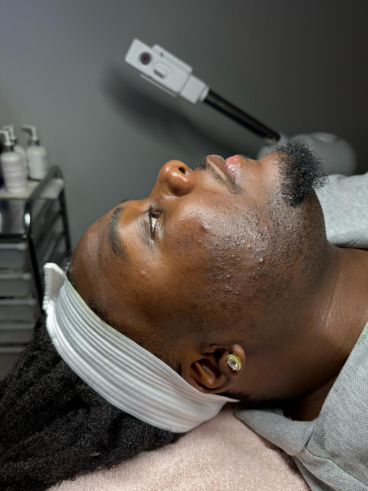 A man lying down with his head tilted back, wearing a white headband and a gray shirt, in a spa or treatment setting with a facial device above his head and skincare bottles in the background.